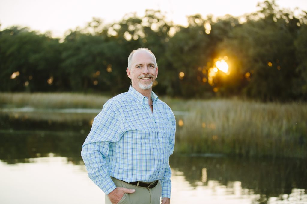 An older man with graying hair and a goatee, wearing a light blue and green plaid button-down shirt and khaki pants, stands smiling in front of a calm body of water with trees in the background during a golden sunset - https://www.rhinspartners.com/