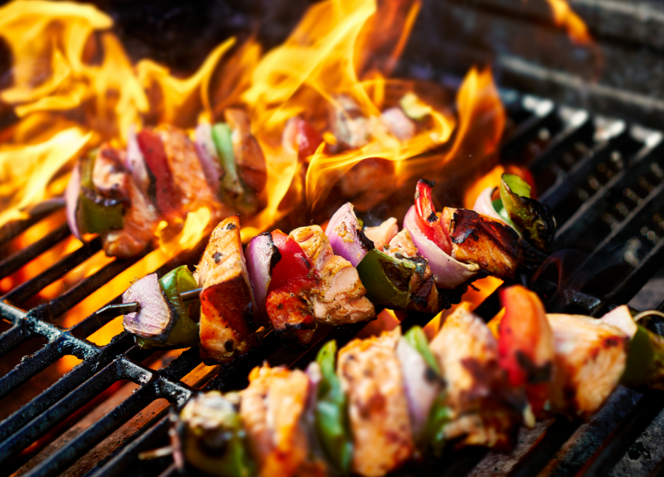 A close-up of chicken and vegetable skewers grilling over bright, orange flames on a black grill grate - https://www.rhinspartners.com/