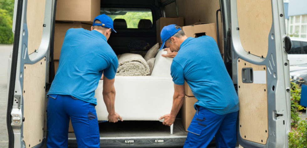 Two professional movers wearing blue uniforms and caps load a white sofa into the back of a moving van filled with cardboard boxes - https://www.rhinspartners.com/