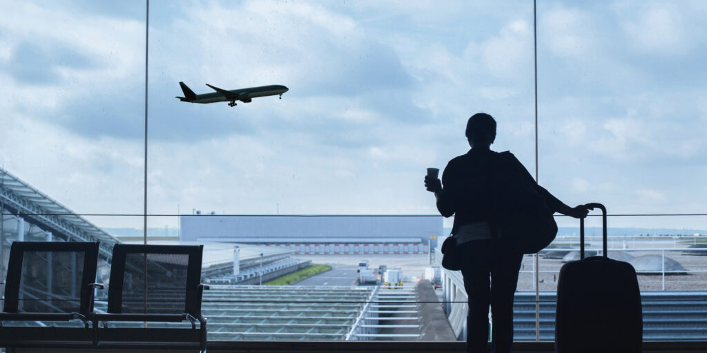 Silhouette of a traveler holding a coffee cup and luggage, looking out of a large airport window at a plane taking off against a cloudy sky - https://www.rhinspartners.com/