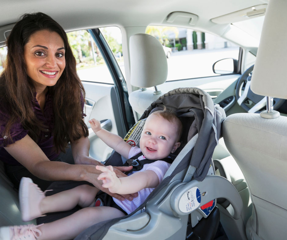 An adult woman with long brown hair smiles at the camera as she secures a smiling infant into a rear-facing car seat in the back of a vehicle - https://www.rhinspartners.com/