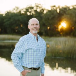 An older man with graying hair and a goatee, wearing a light blue and green plaid button-down shirt and khaki pants, stands smiling in front of a calm body of water with trees in the background during a golden sunset - https://www.rhinspartners.com/